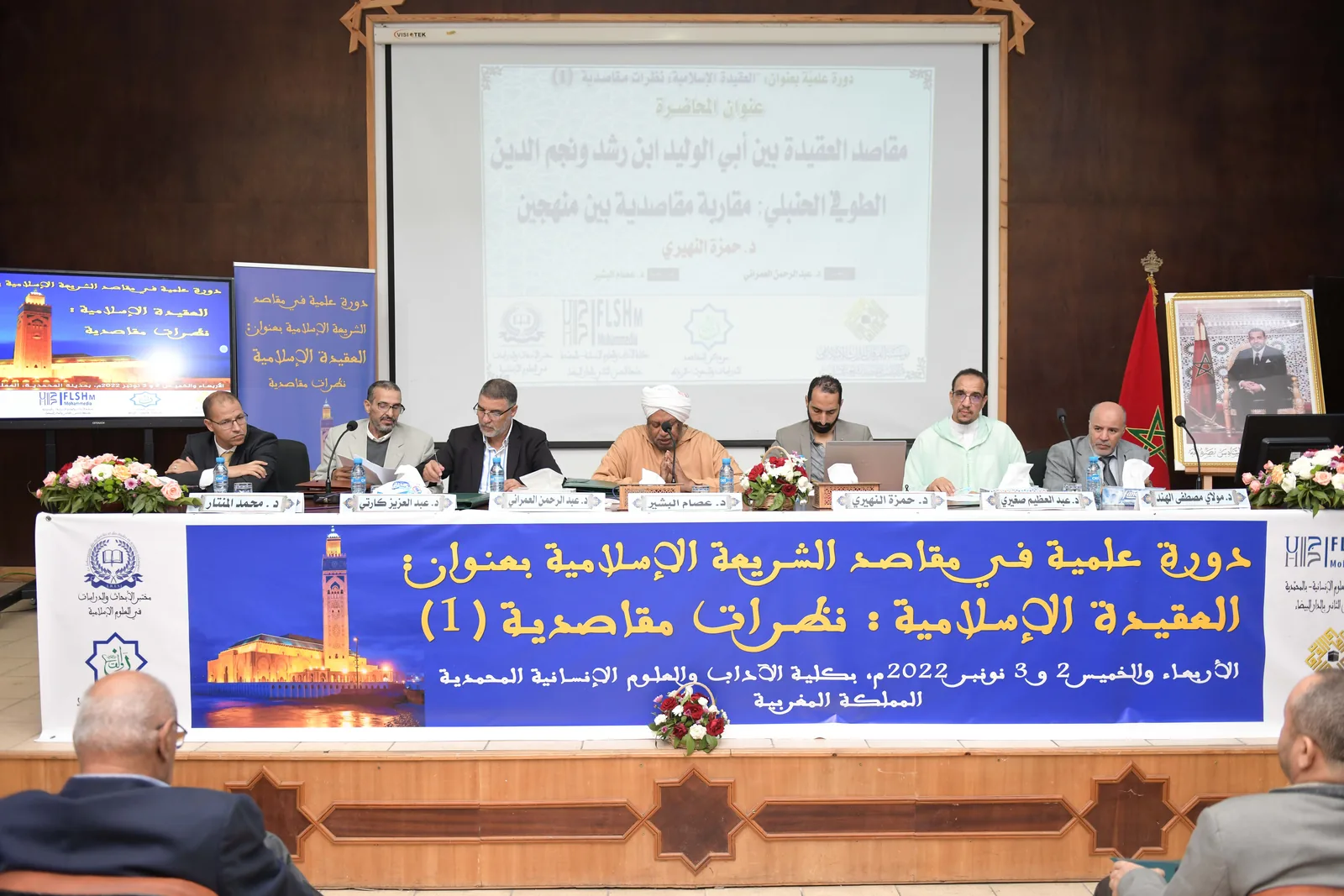 Table ronde de conférence avec écran de projection et public en salle.